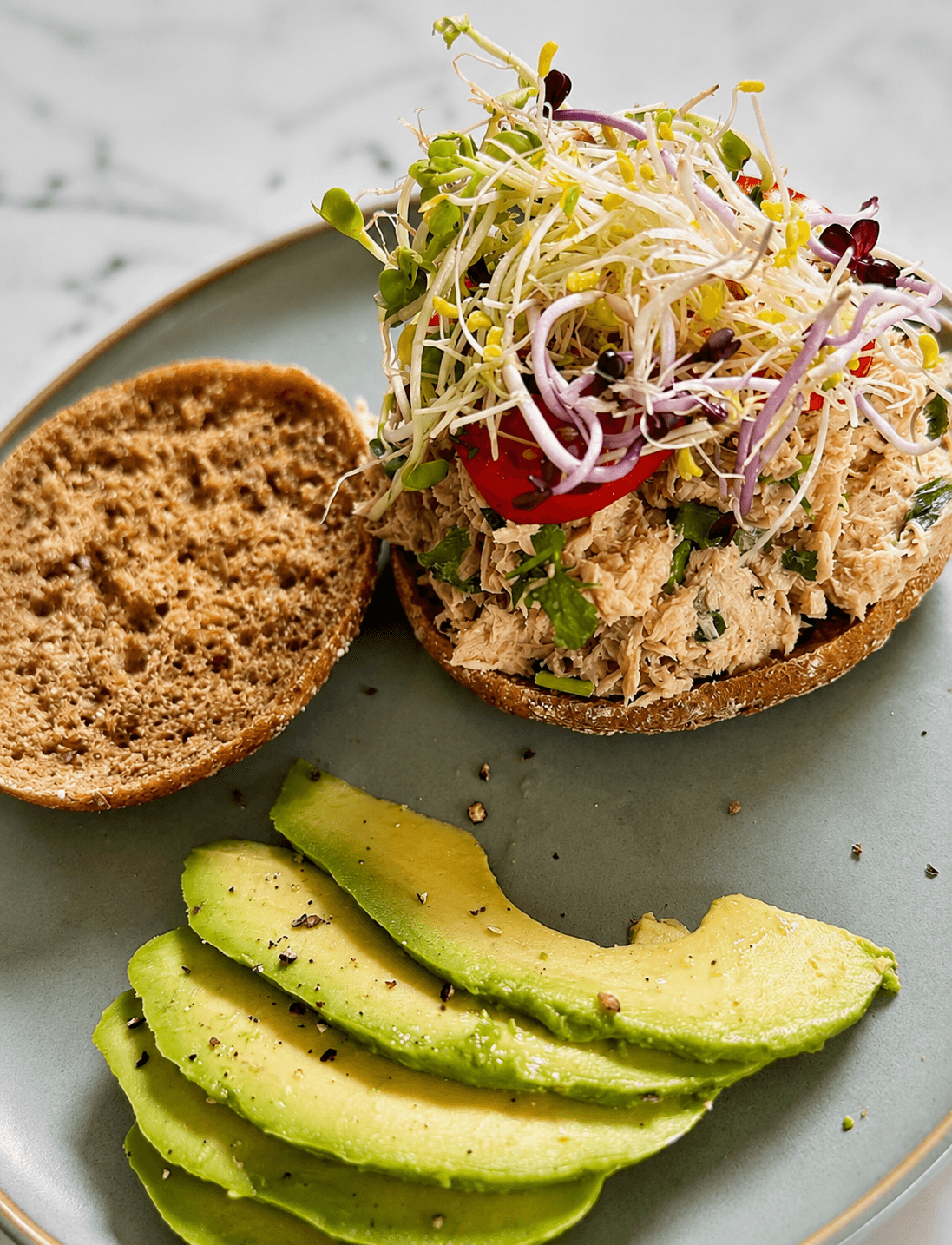 An open-faced tuna melt on toasted sourdough bread topped with tomato slices and a colorful mix of sprouts, served alongside thinly sliced avocado sprinkled with black pepper on a gray plate.