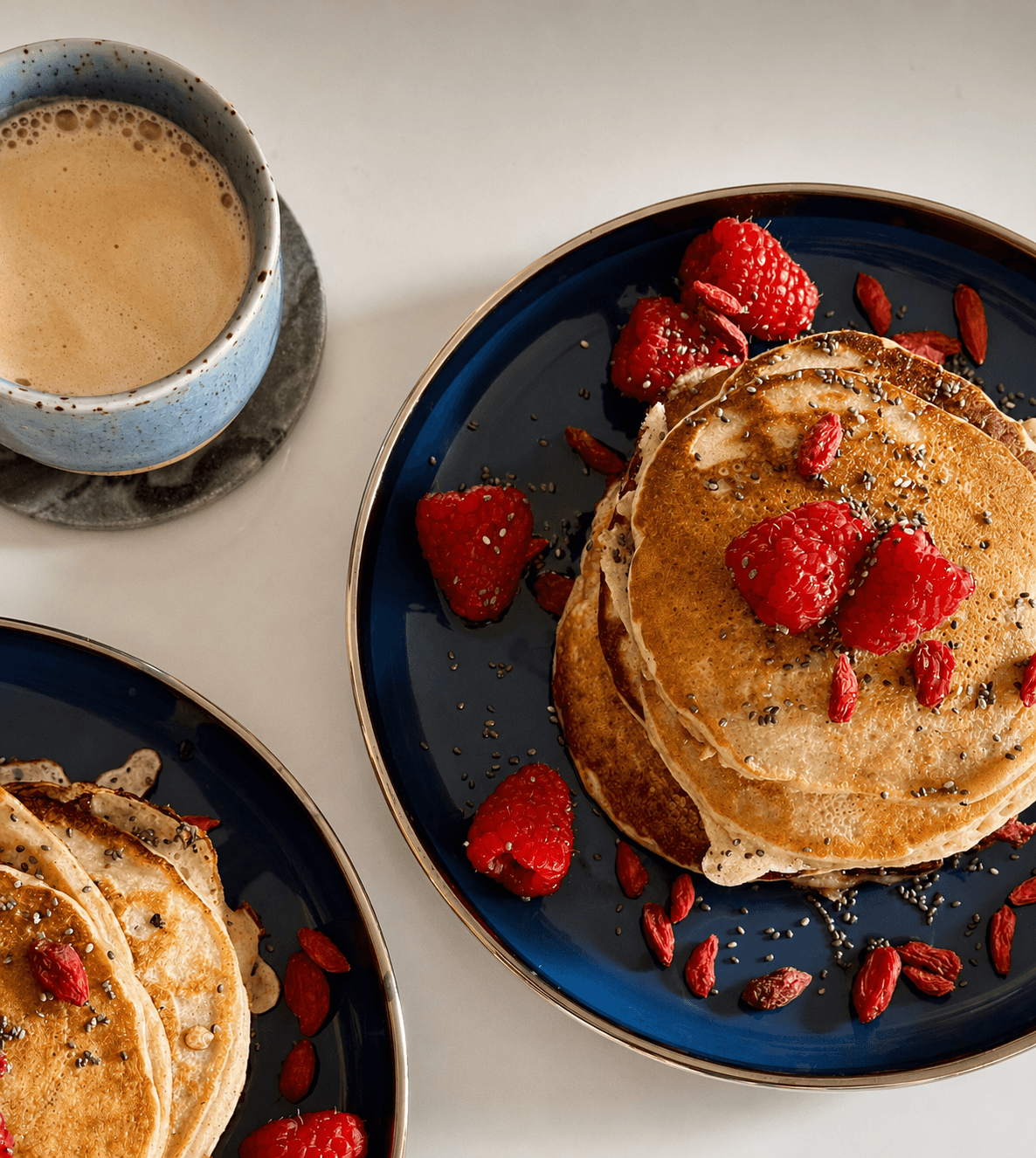 Blue plate with a stack of golden-brown pancakes topped with fresh raspberries, sprinkled chia seeds, and goji berries, next to a blue ceramic cup of coffee with froth on a white surface