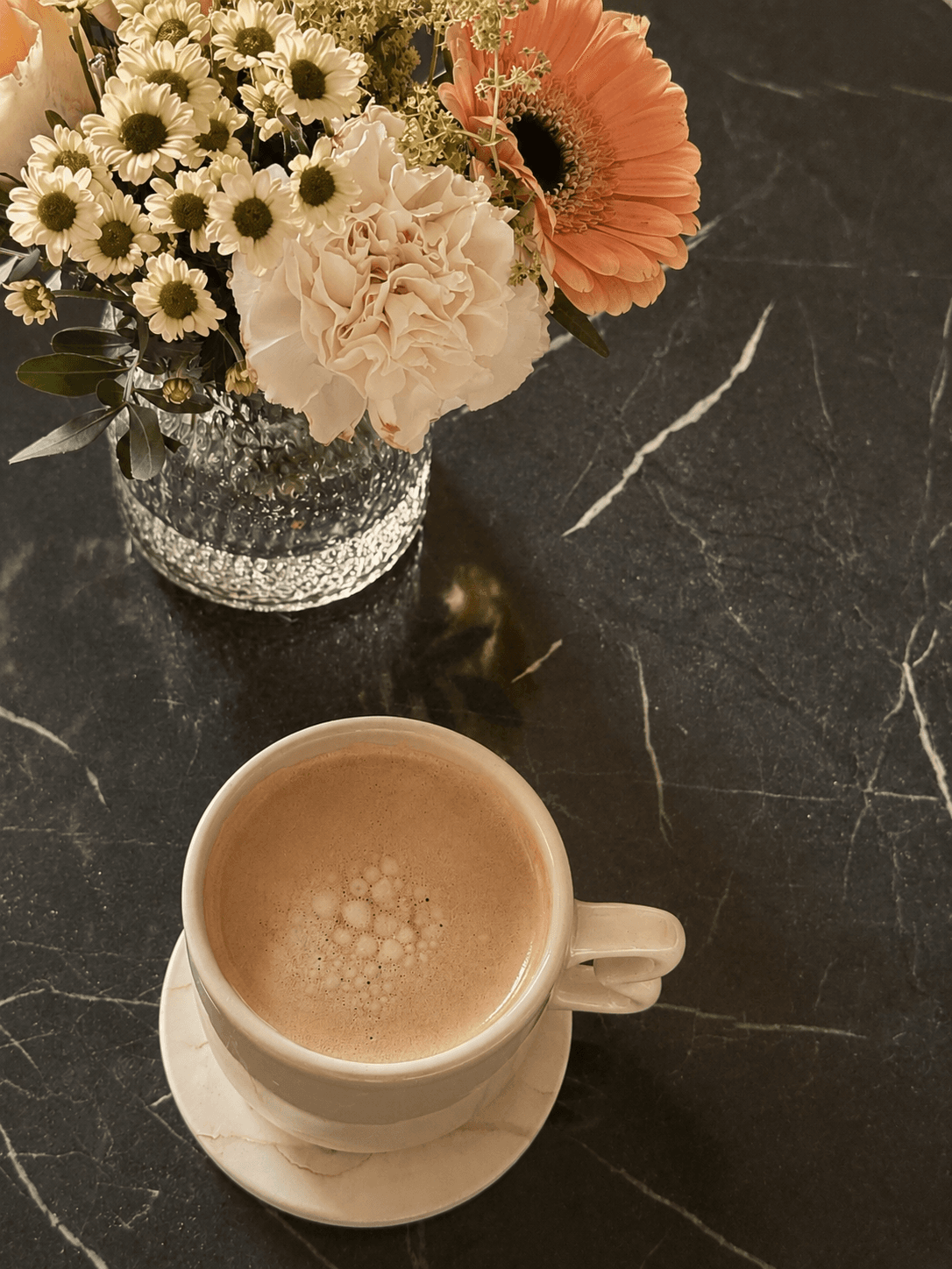 White ceramic cup with a frothy Americano coffee on a marble coaster beside a vase of cream and peach colored flowers, set on a black marble surface