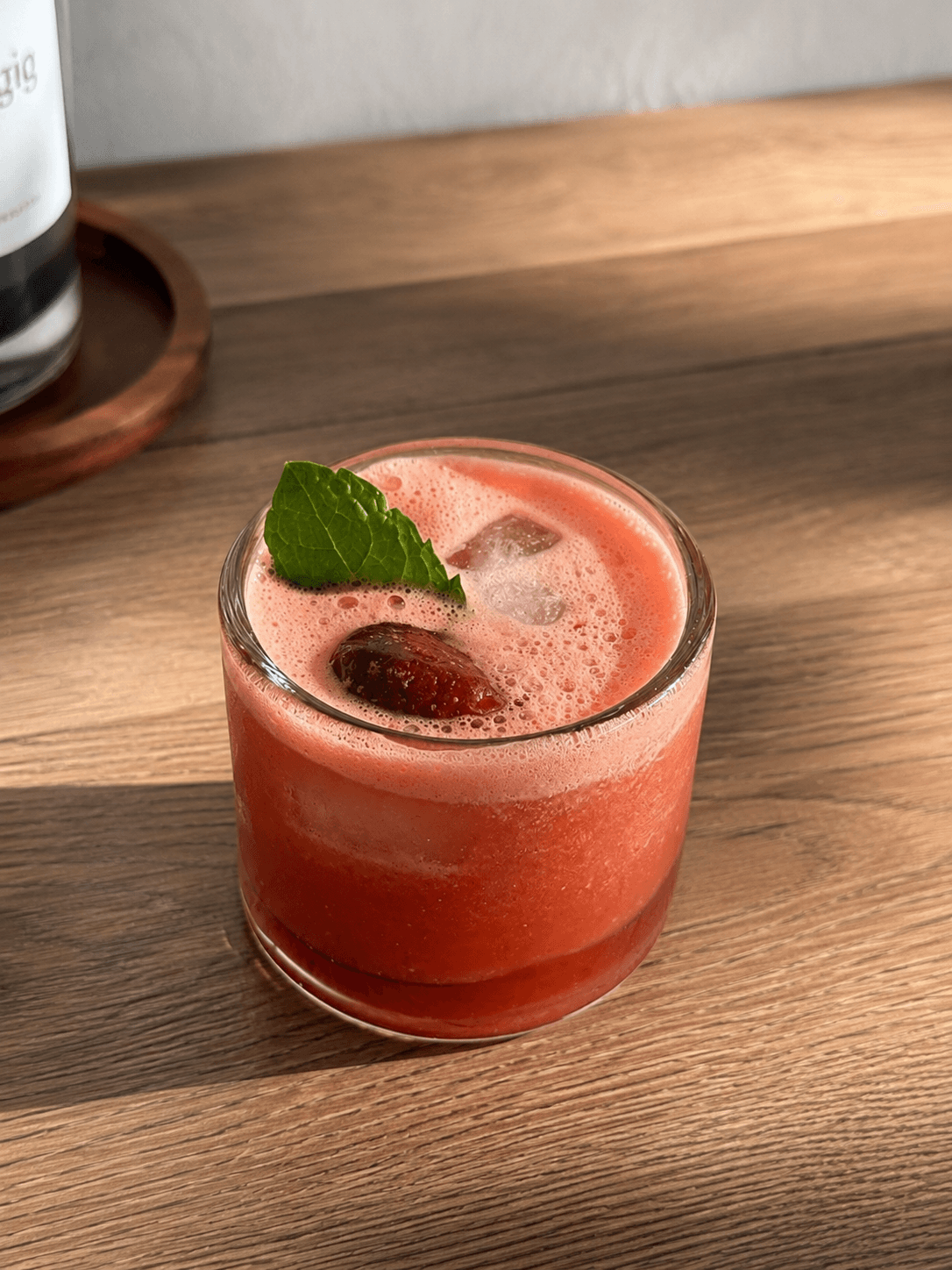 A short glass filled with a pink watermelon drink garnished with a fresh mint leaf and ice cubes, placed on a wooden surface with soft natural light.