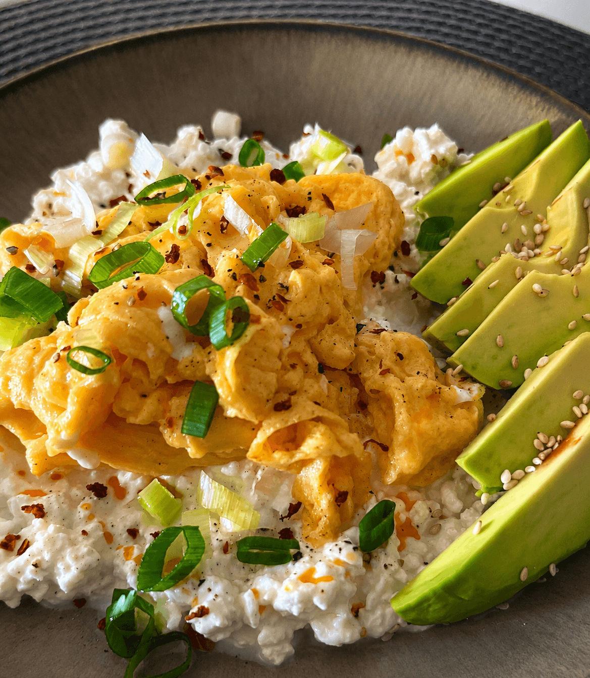 Close-up of a breakfast bowl with scrambled eggs, cottage cheese, sliced avocado sprinkled with sesame seeds, and chopped spring onions with chili flakes on a gray plate