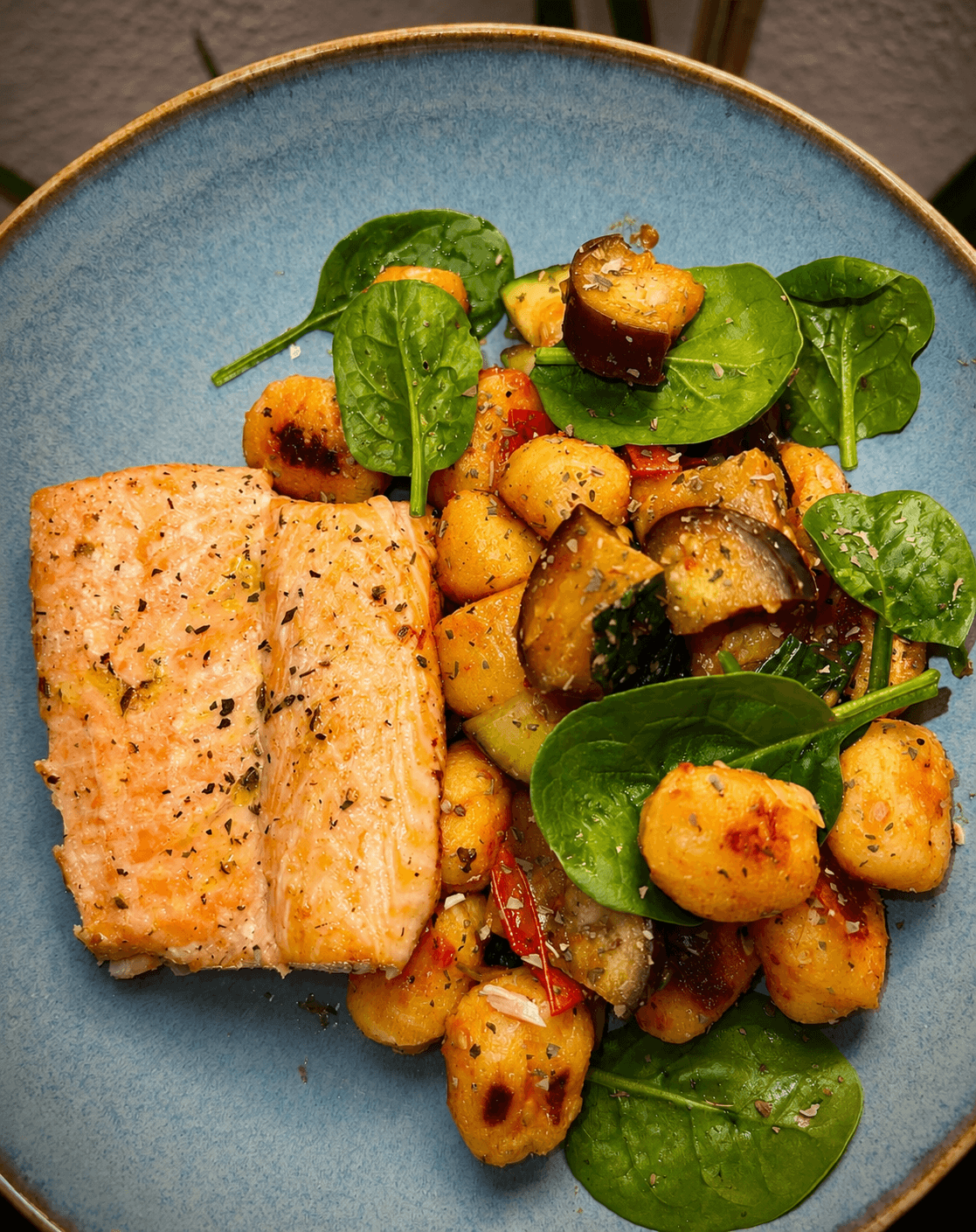 Blue plate with two pieces of seared salmon beside roasted gnocchi and mixed vegetables including spinach leaves, diced zucchini, eggplant, and tomatoes