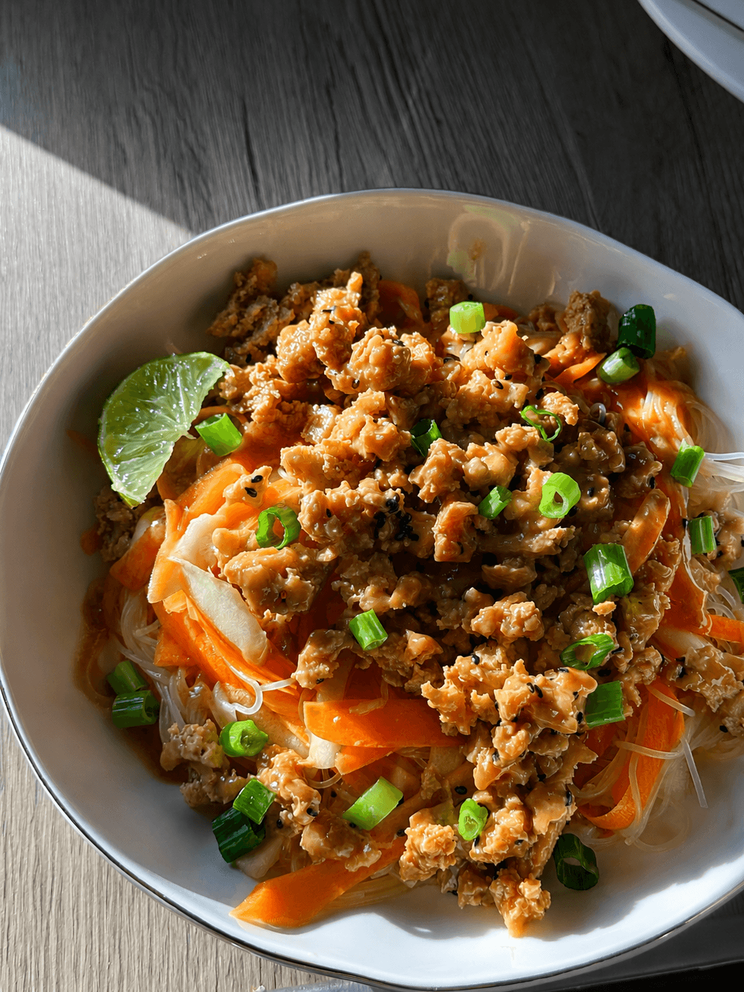 White bowl with a colorful summer-inspired dish featuring crumbled tofu, carrot ribbons, cucumber strips, rice noodles, garnished with green onion slices and a lime wedge, in natural light on a wooden surface