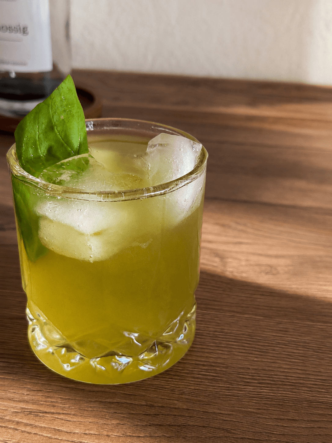 A glass of pale green basil citrus refresher with ice and a fresh basil leaf on top, sitting on a wooden surface.