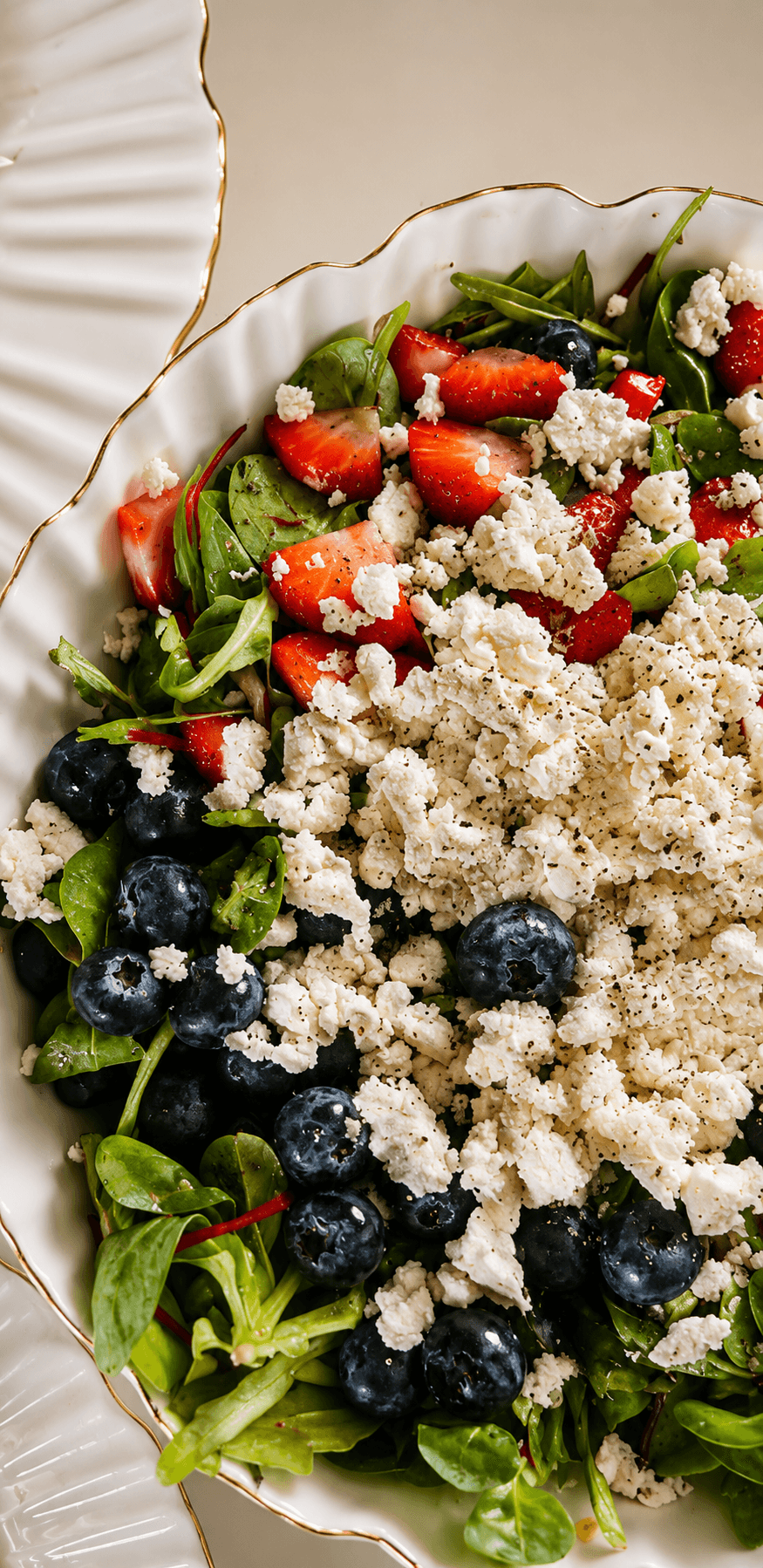 Close-up view of a salad bowl containing mixed greens, blueberries, sliced strawberries, and crumbled feta cheese, lightly drizzled with dressing and sprinkled with black pepper.
