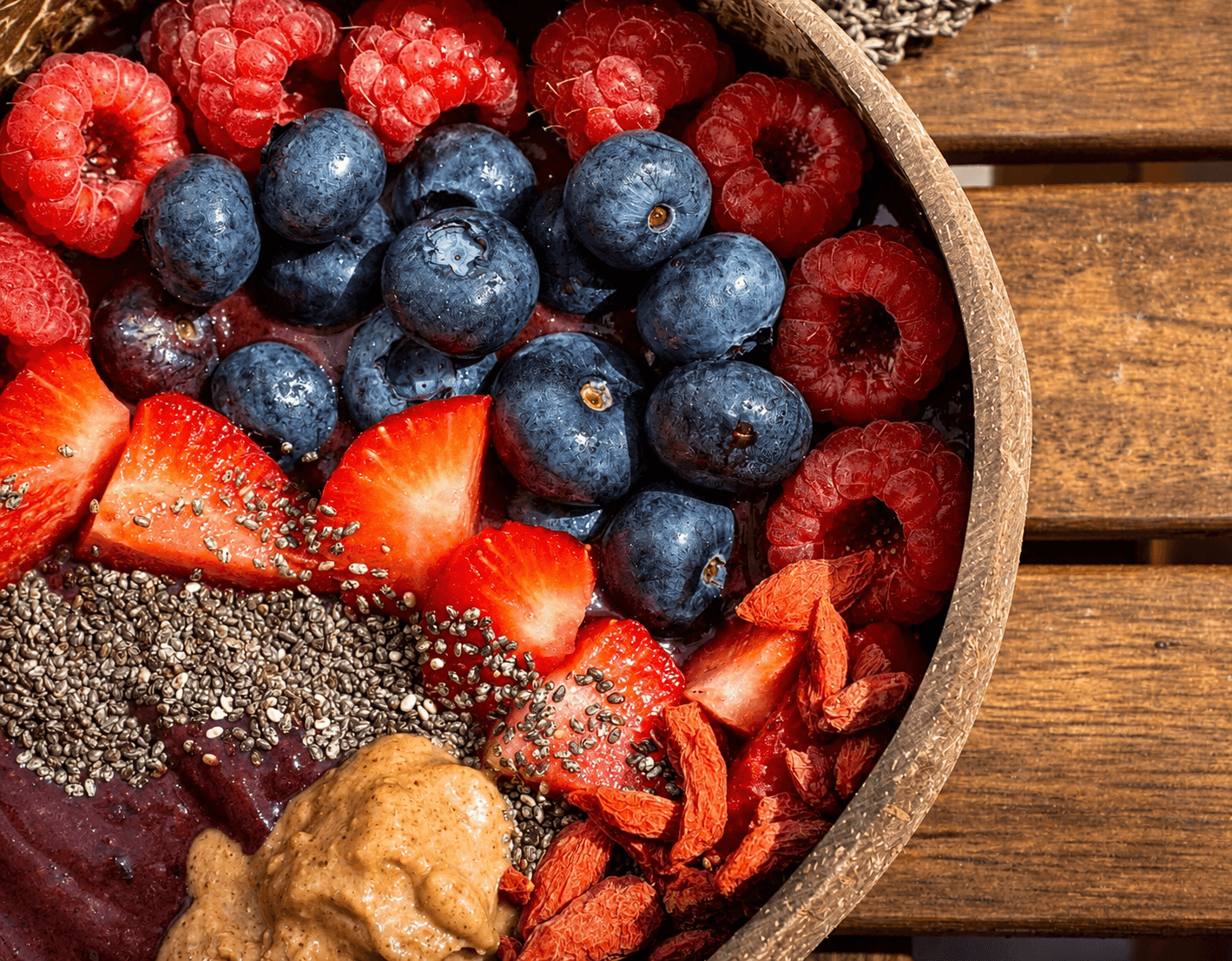 Close-up of a wooden bowl filled with blueberries, raspberries, sliced strawberries, goji berries, chia seeds, peanut butter, and acai puree on a wooden surface