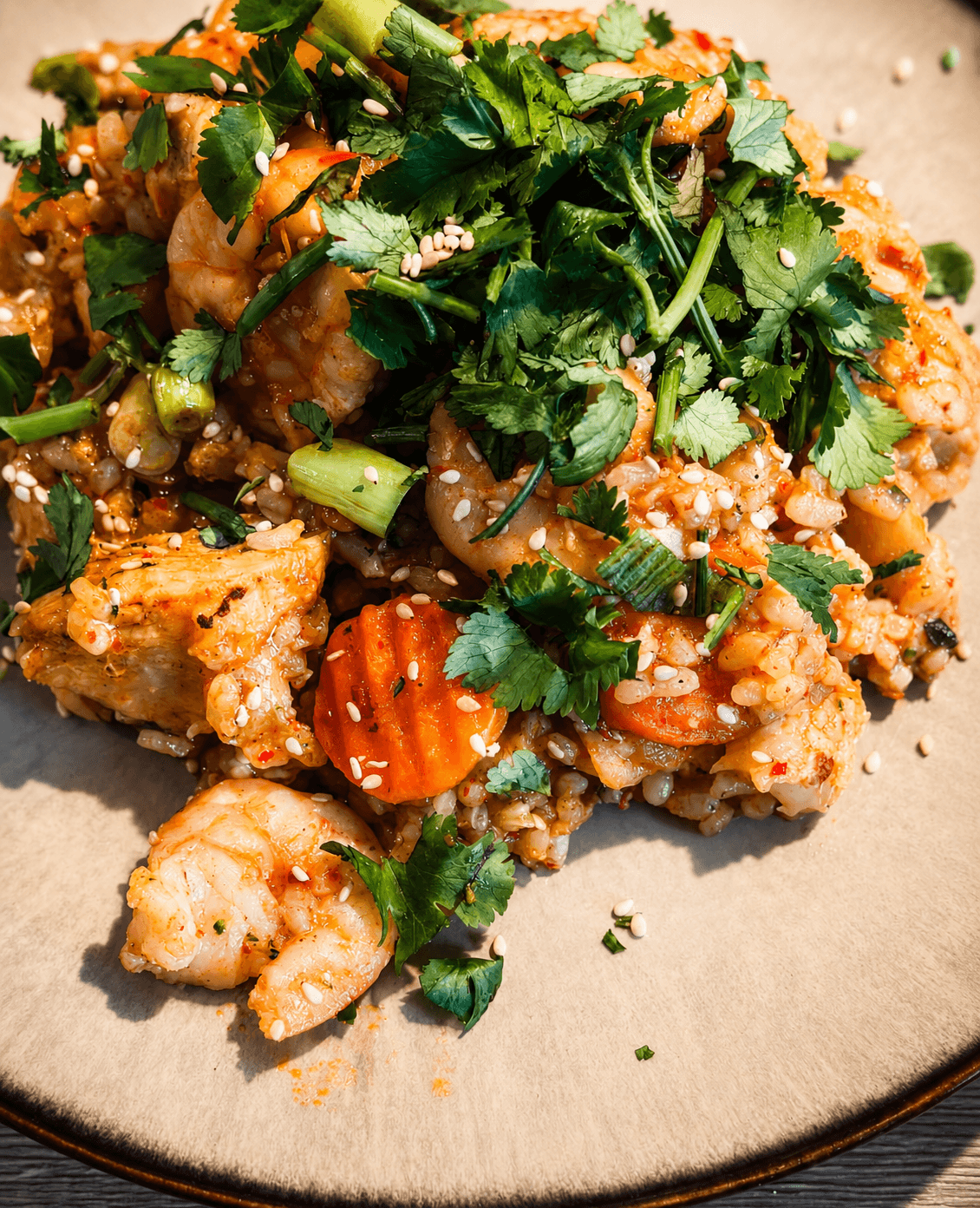 Close-up of kimchi fried rice with cooked prawns, sliced carrots, green onions, cilantro, and sesame seeds on a beige plate