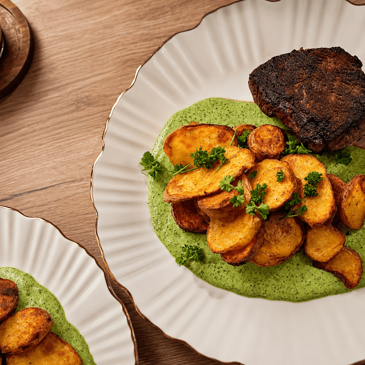 White ceramic plate with air-fried potato slices over green creamy sauce, topped with fresh parsley, alongside a piece of seared steak, all on a wooden table.