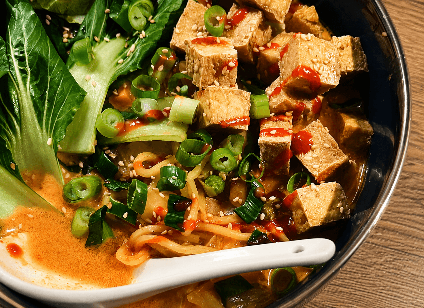 Close-up of a bowl containing ramen noodles, cubed tofu with chili sauce, fresh pak choi leaves, chopped scallions, and sesame seeds in a broth, with a white ceramic spoon resting inside.
