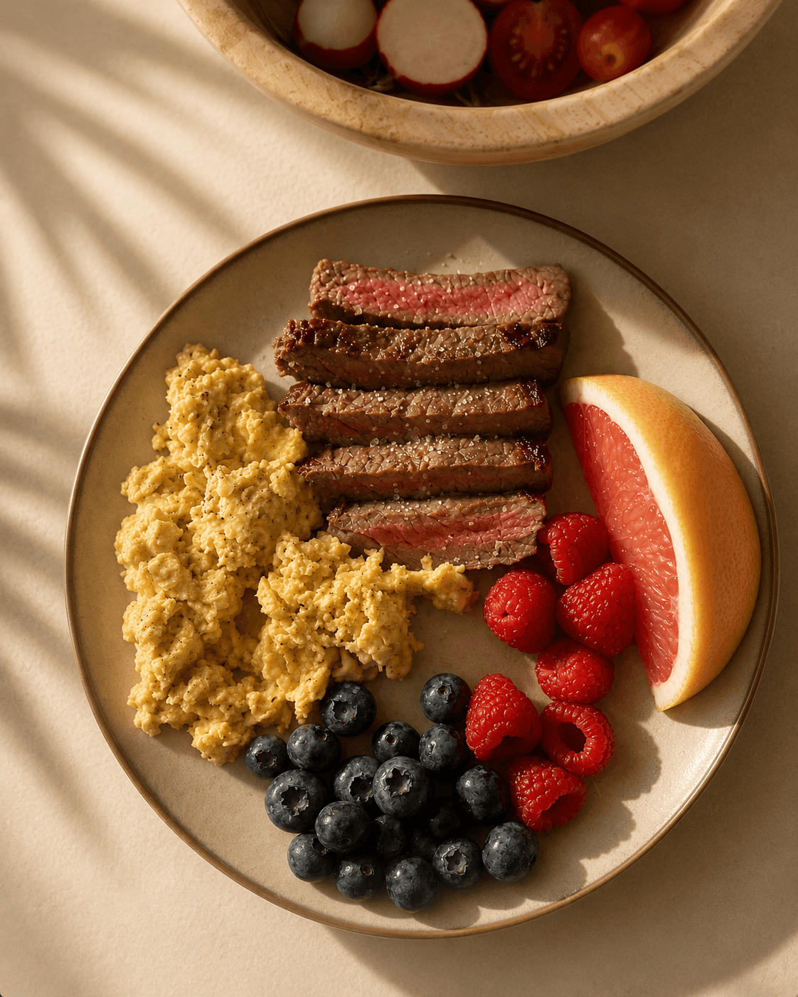 A plate with scrambled eggs, medium-rare steak slices, fresh blueberries, raspberries, and a grapefruit wedge.