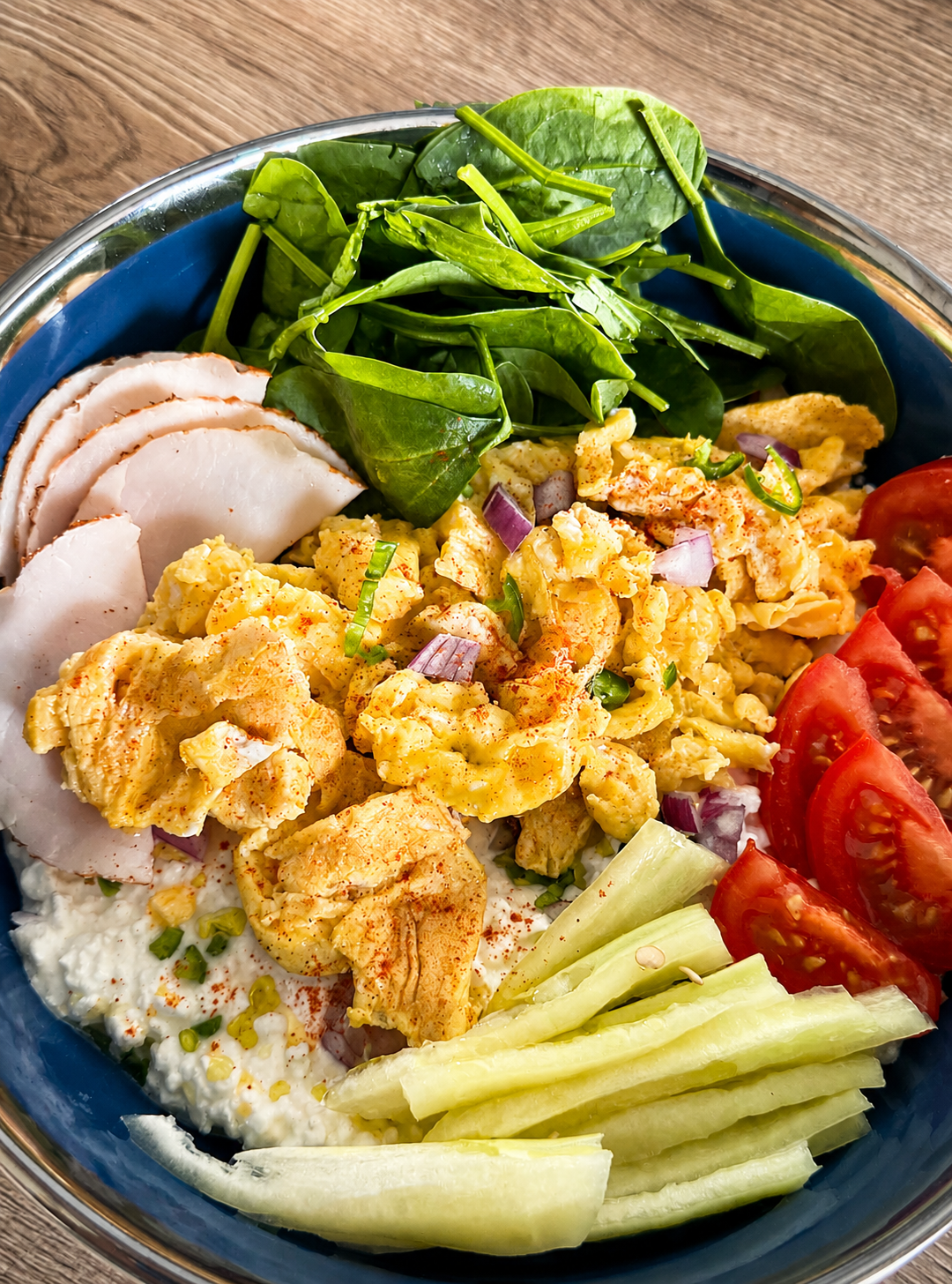 A blue bowl with scrambled eggs, slices of cooked chicken breast, fresh spinach leaves, cottage cheese, sliced cucumber sticks, and tomato wedges arranged neatly on a wooden table