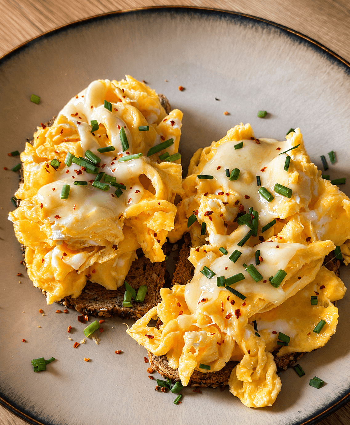 Close-up of two slices of sourdough bread topped with soft scrambled eggs, melted low-fat cheese, chopped chives, and a sprinkle of red pepper flakes on a gray plate