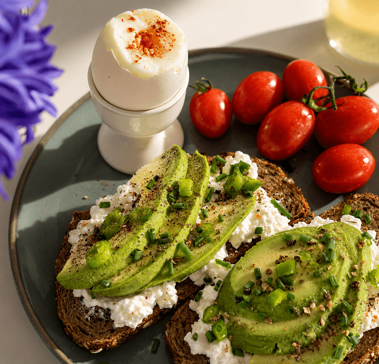 A breakfast plate showing two slices of seeded sourdough topped with cottage cheese, sliced avocado, and chopped fresh chives, accompanied by a soft-boiled egg in an egg cup and a small cluster of ripe cherry tomatoes.