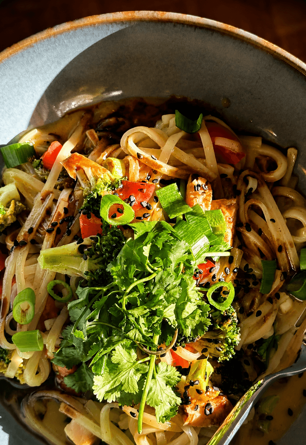A bowl of red curry with chicken, broccoli, red peppers, wheat noodles, topped with fresh cilantro, green onions, and black sesame seeds