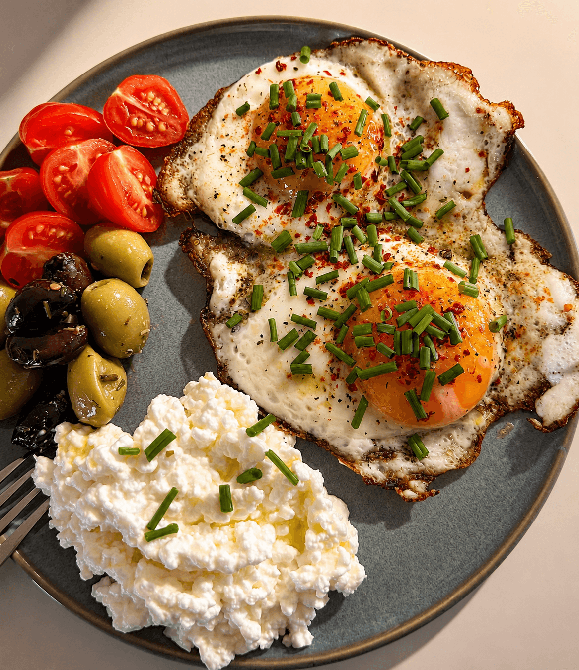 A plate with two sunny-side-up eggs sprinkled with chopped chives, served alongside cottage cheese, halved cherry tomatoes, and a mix of green and black olives.