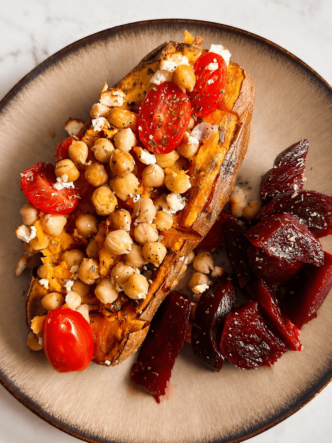 Baked sweet potato topped with chickpeas, halved cherry tomatoes, crumbled feta on a beige plate with sliced roasted red beets on the side