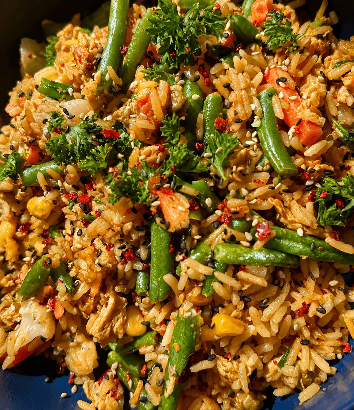 Close-up of vegetable fried rice with green beans, carrots, corn, herbs, and sprinkled sesame seeds in a dark blue bowl