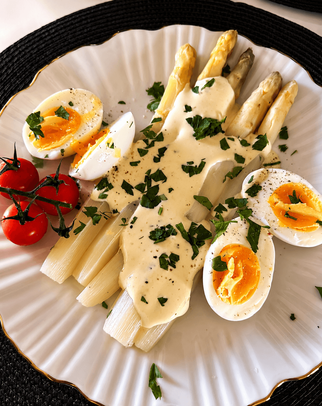 White asparagus spears topped with hollandaise sauce, halved soft-boiled eggs garnished with chopped parsley, and a small vine of cherry tomatoes on a white plate with gold trim