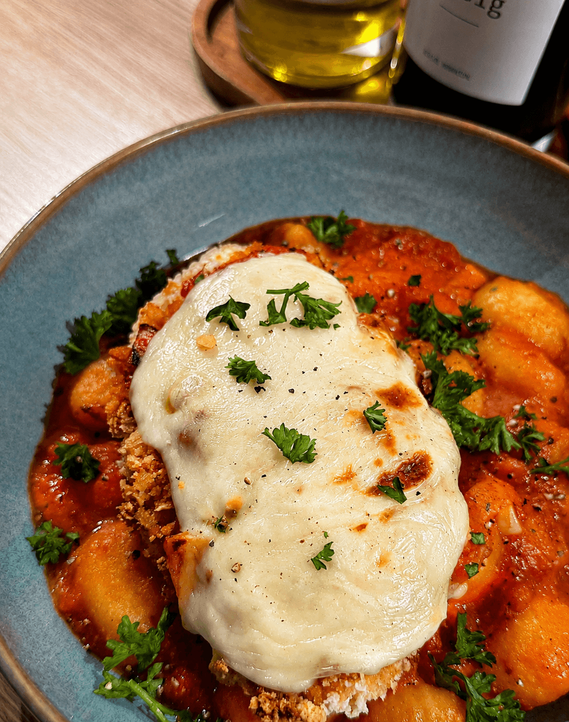 Close-up of a serving of chicken parmesan with melted mozzarella cheese over sweet potato gnocchi topped with fresh parsley in a blue bowl.