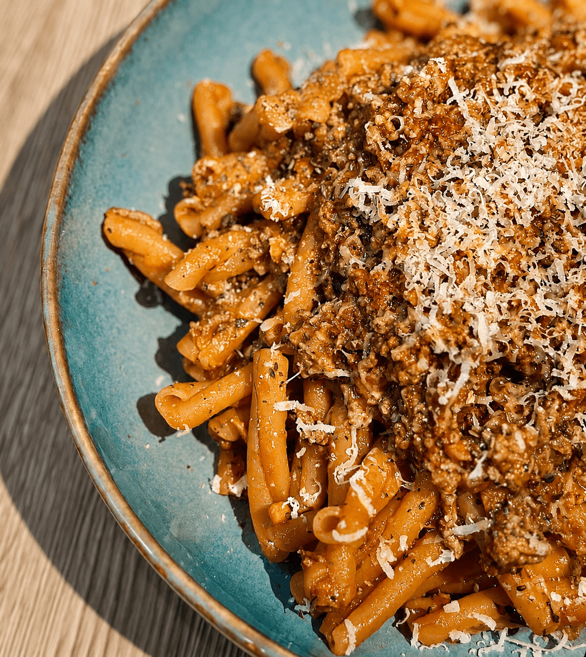 Close-up of a blue bowl filled with red lentil pasta topped with minced beef bolognese and grated cheese on a wooden surface.