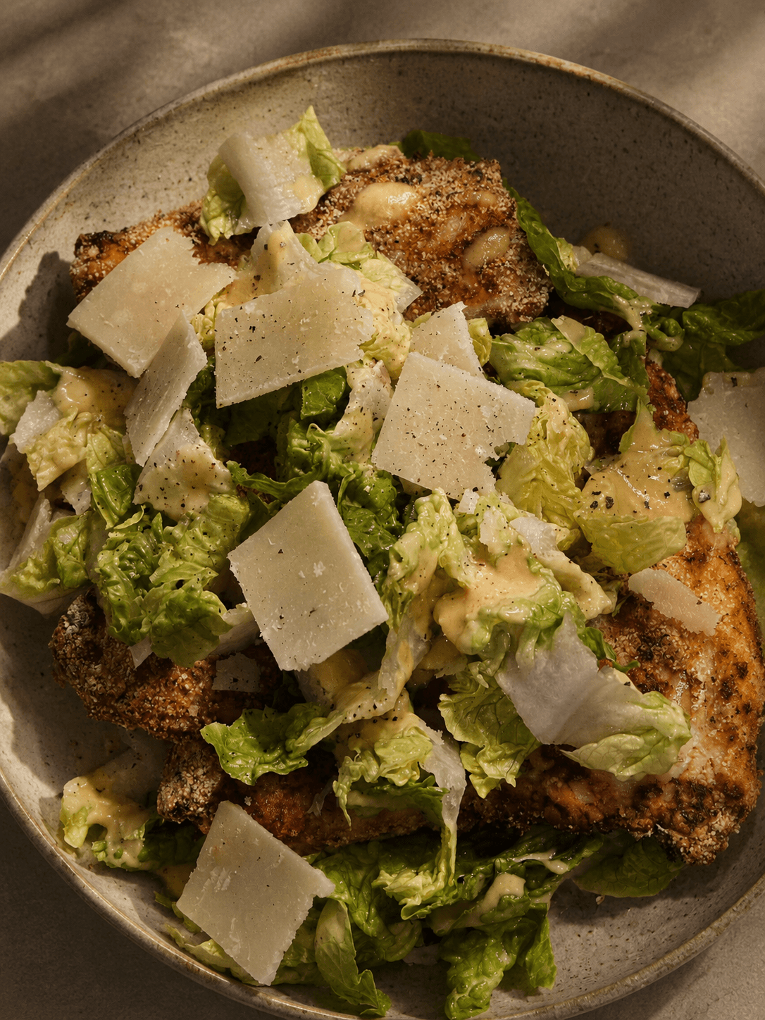 Bowl of Caesar salad with breaded chicken pieces, romaine lettuce, shaved parmesan cheese, and creamy dressing, photographed in soft natural light.