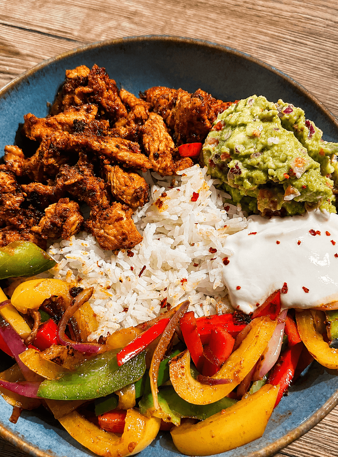 A blue bowl with sliced spiced chicken, sautéed yellow, red, and green bell peppers with onions, white rice, guacamole, and a dollop of yogurt sprinkled with chili flakes.