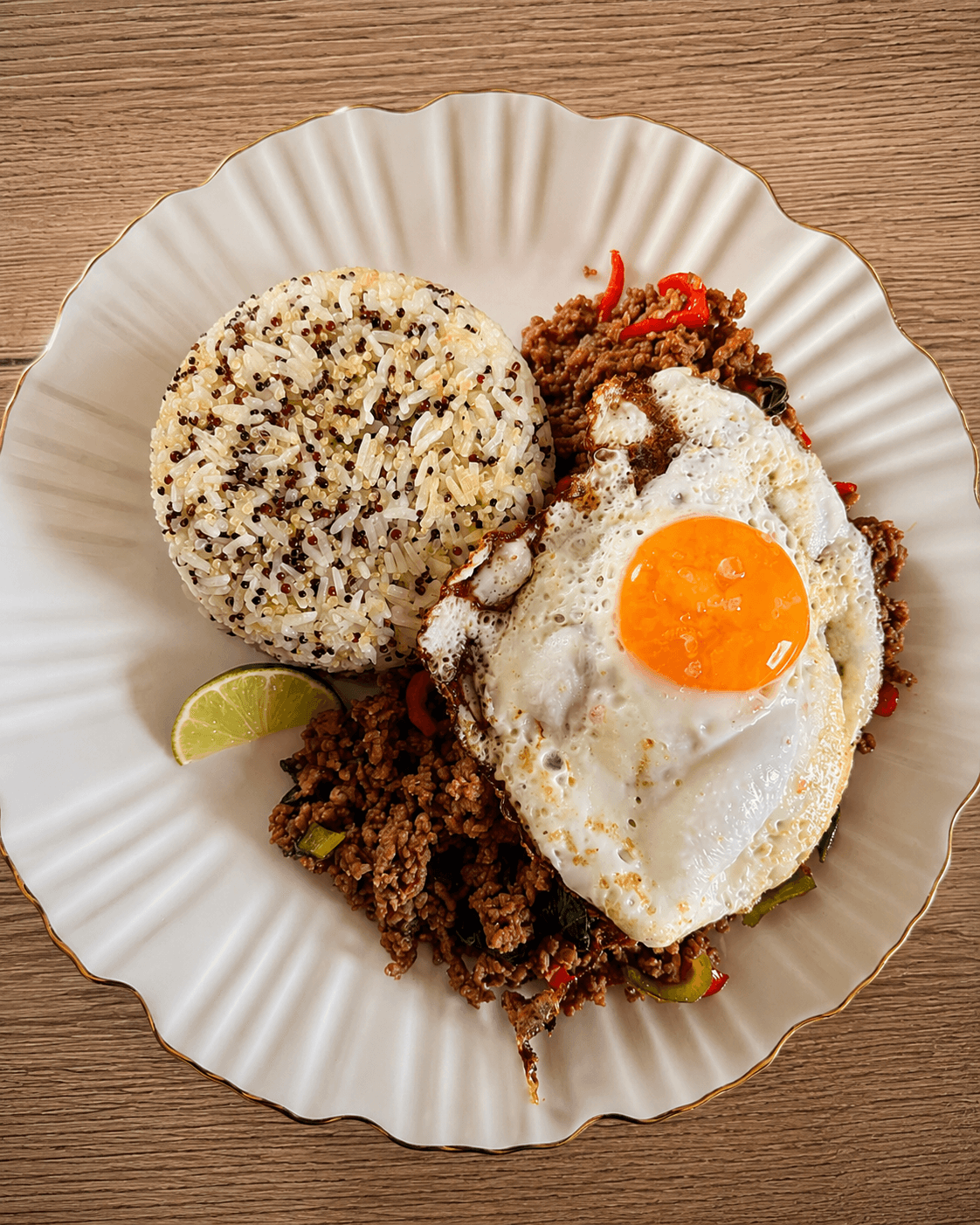 A white scalloped plate holding a portion of mixed jasmine rice and quinoa, a serving of spiced ground meat with red chili slices and basil, topped with a sunny-side-up egg and a lime wedge on the side.